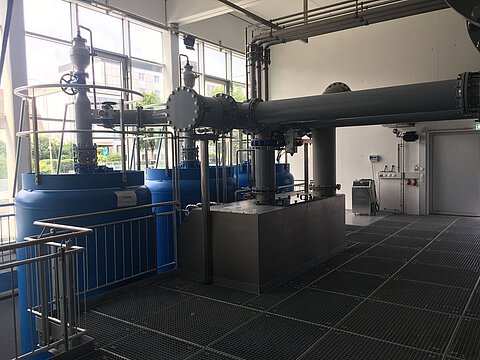 Innenansicht einer technischen Anlage zur Wasseraufbereitung. Zu sehen sind große blaue Behälter, Rohre und verschiedene Armaturen. Das Gelände ist gut beleuchtet und modern gestaltet, mit einem rutschfesten Bodenbelag.