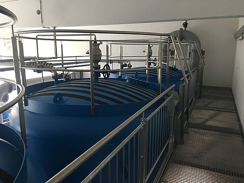 Blick auf mehrere große blaue Tanks in einer industriellen Anlage. Die Tanks sind durch ein Geländer gesichert, und verschiedene Rohre und Ventile sind sichtbar. Der Boden ist mit einem Gitterrost abgedeckt, und es gibt eine Wand im Hintergrund.
