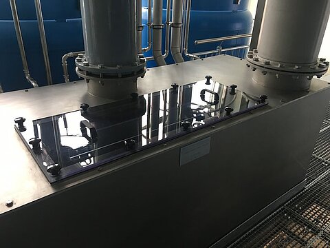 Ein metallischer Tank mit zwei hohen Rohren an der Oberseite. Auf der Tankoberfläche befindet sich eine transparente Abdeckung. Im Hintergrund sind weitere blaue Tanks und Rohrleitungen sichtbar. Die Umgebung wirkt industriell und technisch.