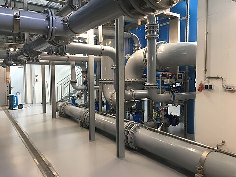 Eine industrielle Rohrleitungsanlage mit großen grauen Rohren, die durch den Raum verlaufen. Der Boden ist glatt und grau, und im Hintergrund sind einige Kontrollgeräte sowie eine Treppe sichtbar. Das Bild zeigt eine hohe technische Ausstattung.