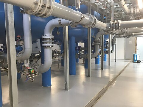 In einer industriellen Umgebung sind große, glänzende Rohre zu sehen, die durch blauen Tanks verlaufen. Der Boden ist aus grauem Material und es gibt verschiedene Ventile und Ventilkästen. Helle Beleuchtung sorgt für eine klare Sicht auf die Ausstattung.