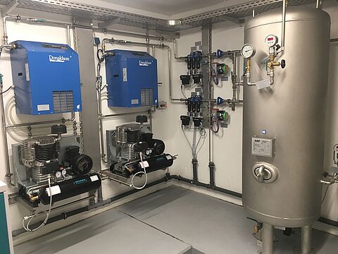 Der Raum zeigt eine technische Anlage mit zwei Kompressoren und einem größeren Druckbehälter. Die Geräte sind an Rohren und Ventilen angeschlossen, die an den Wänden montiert sind. Die Wände und der Boden sind neutral gehalten, um die Umgebung zu betonen.