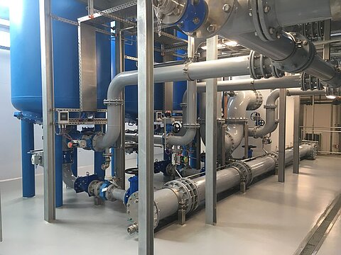 Die Abbildung zeigt eine industrielle Anlage mit blauen Wassertanks und zahlreichen Rohren, die in verschiedenen Richtungen verlaufen. Die Rohre sind aus Metall und haben mehrere Ventile zur Steuerung des Flusses. Der Boden ist glatt und hell, und die Wände sind neutral gehalten.