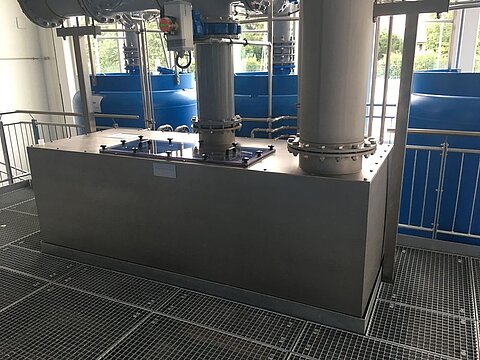 Industrieanlage mit großen, silbernen Rohren und einem rechteckigen Metallbehälter auf einer Gitterplattform. Im Hintergrund sind blaue Wassertanks sichtbar. Die Szene vermittelt einen Eindruck von technischer Infrastruktur und Verarbeitung.