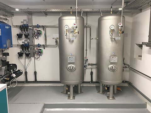 Zwei große, runde Silberbehälter stehen nebeneinander in einem technischen Raum. Sie sind mit Rohren und Messgeräten verbunden. An der Wand sind weitere technische Geräte und Steuerungen angebracht. Der Boden ist grau und die Wände sind schlicht.