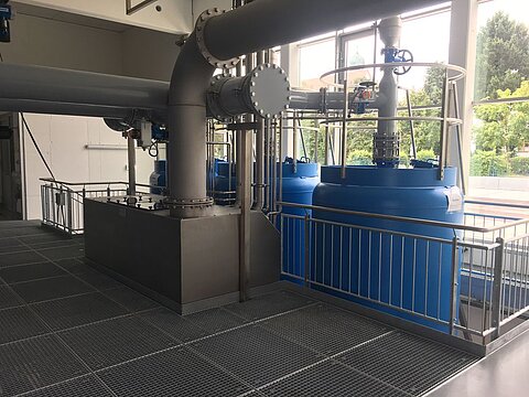 Industrielle Anlage mit blauen Tanks und großen Rohren. Die Tanks stehen auf einem Gitterboden, umgeben von einem Geländer. Helle, moderne Umgebung mit Fenstern, durch die Tageslicht strömt.