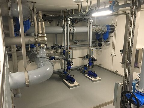 Ein technischer Raum mit Rohrleitungen und Ventilen. Sichtbare blaue Ventile und Rohre verlaufen durch den Raum, ergänzt durch eine große graue Pipeline. Der Boden ist grau und gut beleuchtet. Es gibt auch einige technische Geräte an den Wänden.