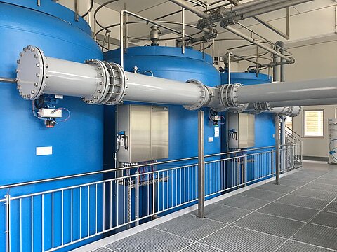 Drei große, blaue Tanks stehen nebeneinander in einem industriellen Raum. Sie sind an Rohre angeschlossen, die durch den Raum führen. Eine Metallgeländer umschließt die Fläche, auf der sich die Tanks befinden.