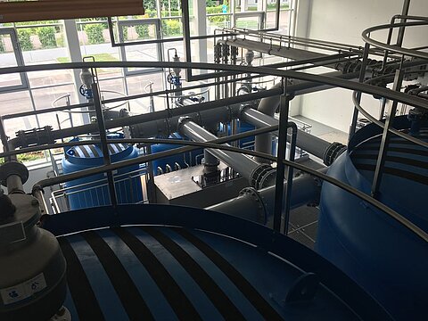 Blick auf eine Industrieanlage mit blauen Tanks und sichtbaren Rohren. Die Struktur zeigt verschiedene Leitungen und Ventile in einem modernen, gut beleuchteten Raum.