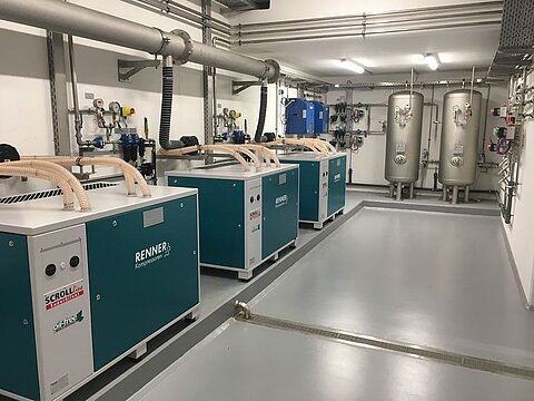 Eine technische Umgebung mit mehreren Kompressoren in blauer und grauer Ausführung. Sie sind auf einem grauen Boden aufgestellt, mit sichtbaren Rohrleitungen und verschiedenen Steuerungs- und Messgeräten an den Wänden. Die Beleuchtung ist hell und gleichmäßig.