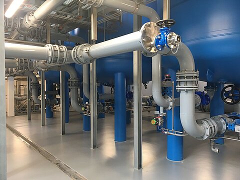Industrielle Umgebung mit großen blauen Wassertanks und zahlreichen Rohrleitungen, die durch verschiedene Ventile verbunden sind. Die Rohre verlaufen entlang des Bodens und sind an Metallstützen montiert. Die Bodenfläche ist eben und hell.