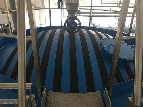 Ein runder Behälter mit blauen und schwarzen Streifen, der sich im Innenbereich einer Industrieanlage befindet. Im oberen Teil ist ein Ventil sichtbar, und Treppenführung aus Metall führt zu dessen Zugang. Die Umgebung zeigt große Fenster und Metallstrukturen.