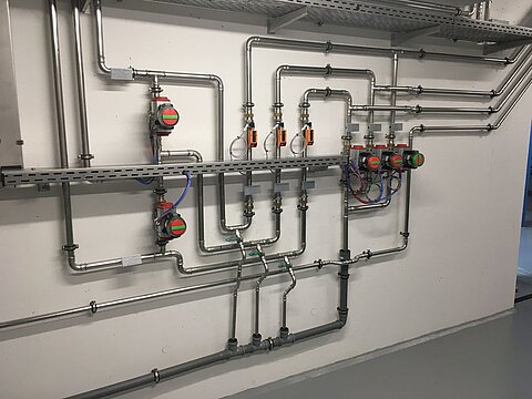 Eine Wand mit komplexem Rohrsystem aus Metallrohren, Ventilen und Messgeräten. Die Rohre verlaufen in verschiedenen Richtungen, verbunden durch Anschlussstücke und Dichtungen. Einige Geräte sind farblich gekennzeichnet. Der Boden ist gepflastert und die Wand ist schlicht gehalten.