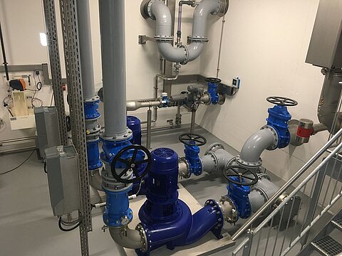 Das Bild zeigt eine technische Anlage mit blauen Ventilen, Rohren und Pumpen in einem Raum. Die Rohre sind sowohl vertikal als auch horizontal angeordnet, und einige Ventile sind sichtbar. Der Boden ist grau und die Wände sind weiß. Es handelt sich um eine industrielle Umgebung.