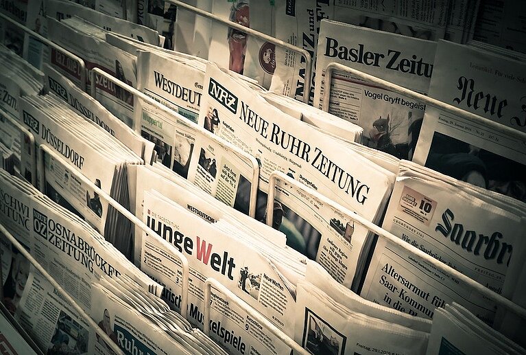 Eine Sammlung von Zeitungen liegt in einem Zeitungsständer. Die Titelblätter sind teils sichtbar, darunter verschiedene deutschsprachige Zeitungen wie „Basler Zeitung“ und „Neue Ruhr Zeitung“. Die Zeitungen sind dicht nebeneinander angeordnet.