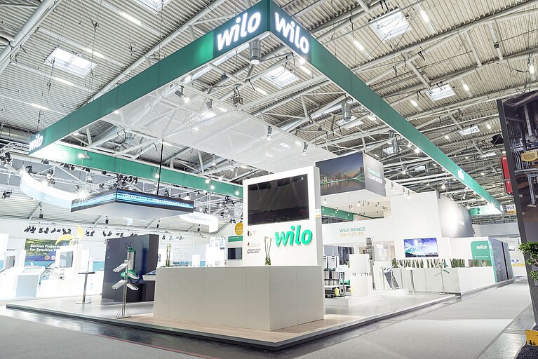 Ein moderner Messestand mit dem Schriftzug "Wilo". Der Stand ist hell und geräumig, mit verschiedenen Displays und Ausstellungsräumen. Das Design ist clean und professionell, mit einer durchgehend weißen und grünen Farbgebung.