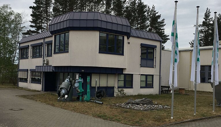 Moderne, zweigeschossige Gebäude mit einer runden Dachkonstruktion. Vor dem Gebäude stehen mehrere Flaggen und es gibt Rasenflächen mit Bäumen im Hintergrund. Der Gehweg vor dem Gebäude ist aus Pflastersteinen.