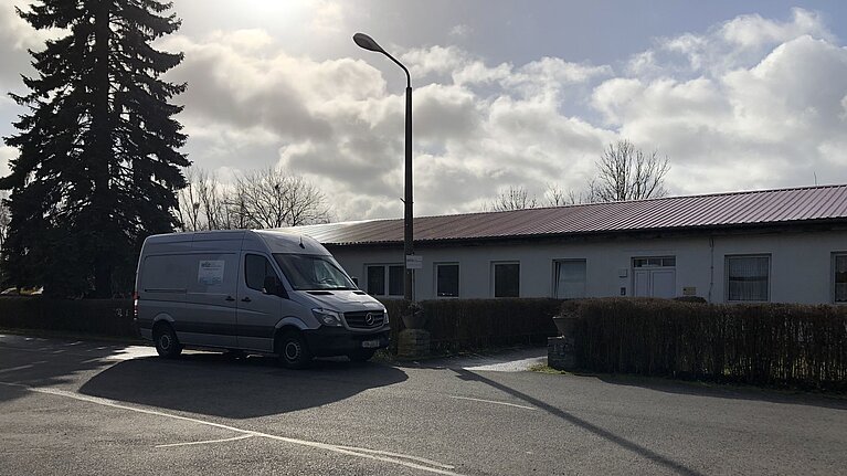 Ein weißer Lieferwagen steht auf einem Parkplatz vor einem flachen Gebäude mit Fenster. Im Hintergrund sind einige Bäume sichtbar, und der Himmel ist bewölkt, was auf wechselhaftes Wetter hindeutet.