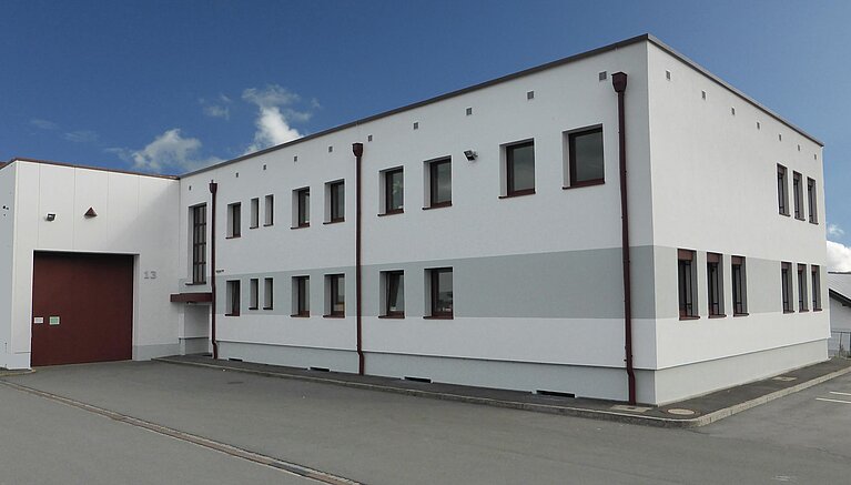 Ein modernes, zweigeschossiges Gebäude mit weißer Fassade und roten Fensterrahmen. Es hat mehrere Fenster und ein großes Eingangstor. Der Himmel ist teilweise bewölkt und es gibt einen Asphaltparkplatz vor dem Gebäude.