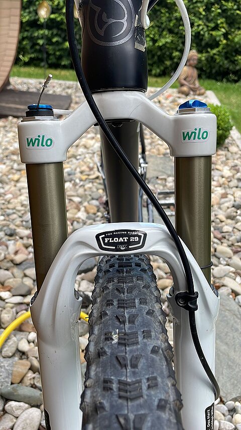 Nahaufnahme einer Mountainbike-Gabel mit einem weißen und grünen Design. Die beiden Federgabelrohre sind in einem goldenen Farbton gehalten. Der Reifen mit grobem Profil ist im unteren Bildbereich sichtbar. Einige Steine liegen im Hintergrund, und der Garten ist unscharf.