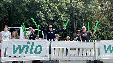 Eine Gruppe von Menschen steht hinter einer Absperrung und hält grüne Lichtröhren in die Luft. Sie tragen T-Shirts mit dem Logo "Wilo". Im Hintergrund sind Bäume zu sehen.