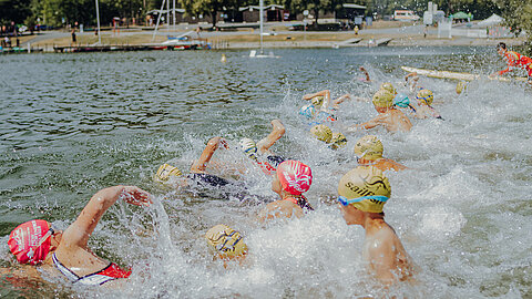Eine Gruppe von Schwimmern in farbigen Badekappen schwimmt im Wasser. Spritzendes Wasser ist zu sehen, während die Schwimmer verschiedene Schwimmstile anwenden. Im Hintergrund sind Bäume und eine ruhige Landschaft.