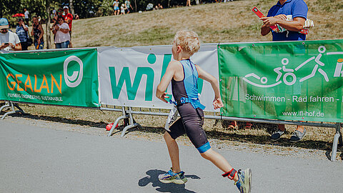 Ein Kind in Sportbekleidung läuft bei einem Wettkampf vorbei. Im Hintergrund sind Banner mit Sponsoren und eine Zuschauergruppe zu sehen. Die Szene vermittelt eine energiegeladene Atmosphäre eines Sportevents.