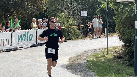 Ein Läufer in einem schwarzen T-Shirt mit dem Logo "wilo" läuft auf einem Weg, während Zuschauer ihn anfeuern. Im Hintergrund sind Teilnehmer und Zuschauer zu sehen. Eine Wegweiser zeigt die Richtung zum "Steinhaus".
