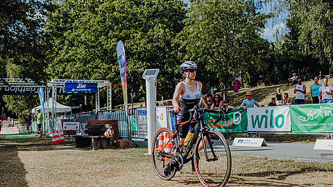 Eine Triathletin mit einem Fahrrad kommt ins Ziel. Sie trägt einen Neoprenanzug und einen Helm. Im Hintergrund sind Zuschauer und Eventbanner zu sehen, die eine sportliche Atmosphäre schaffen.