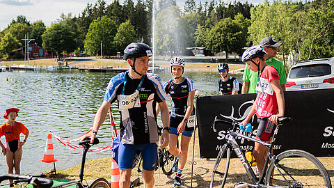 Vier Sportler in Radfahrer-Outfits, einige mit Helmen, bereiten sich am Ufer eines Sees auf ein Rennen vor. Im Hintergrund sind Bäume und eine Wasserfontäne zu sehen. Einige Personen stehen am Rand, während ein Teilnehmer sein Fahrrad in der Hand hält.
