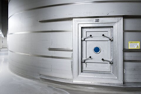 Eine große silberne Metalltür ist an einer runden silbernen Wand angebracht. Die Tür hat eine runde blaue Öffnung in der Mitte und vier Schlösser oder Verschlüsse. Neben der Tür ist ein Warnschild befestigt.