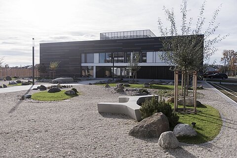 Moderne Architektur eines Gebäudes mit großen Fenstern, umgeben von einem gepflegten Garten mit Kies, Steinen, Rasenflächen und kleinen Bäumen. Der Himmel ist bewölkt, was eine ruhige Atmosphäre schafft.