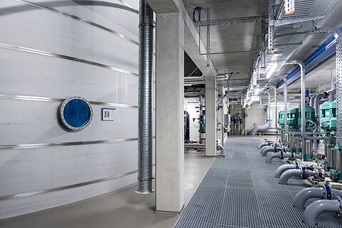 Innenansicht einer industriellen Anlage mit silbernen Wassertanks, einer blauen Zugangsluke und grünen Pumpen. Der Boden ist aus Gitterrost, und es verlaufen Rohre an den Wänden. Die Umgebung wirkt sauber und modern, mit einem hellen und übersichtlichen Design.