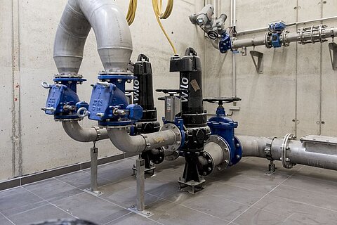 Zwei Pumpen mit blauen Ventilen und Rohren sind in einem technischen Raum installiert. Die Wände sind aus Beton, und der Boden ist gefliest. Die Rohre verlaufen über und neben den Pumpen, die elektrisch betrieben werden.