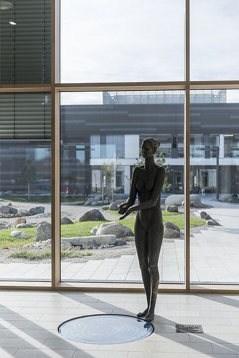 Eine schwarze Statue einer weiblichen Figur steht in einem hellen Raum mit großen Fenstern. Im Hintergrund sind moderne Gebäude und Landschaftsgestaltung mit Steinen und Gras zu sehen.