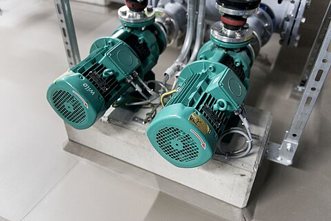 Zwei grüne Elektromotoren stehen auf einem Betonfundament. Sie sind Teil einer Rohrleitung und werden durch verschiedene Anschlüsse mit Rohren verbunden. Der Boden ist grau und eben.