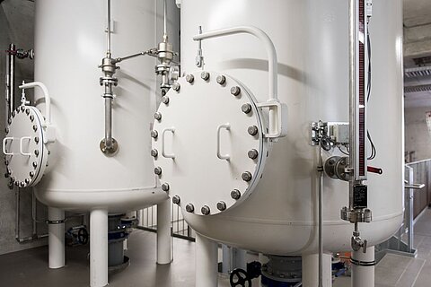 Zwei große, weiße Druckbehälter stehen in einem Industrieumfeld. Jeder Behälter hat eine runde Tür mit vielen Schrauben und ist mit Rohren und Ventilen verbunden. Im Hintergrund sind verschiedene Industrieelemente sichtbar, die auf eine technische Umgebung hinweisen.
