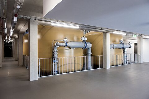 Zwei große, gelbe Tanks stehen in einem modernen Raum. Sie sind durch Metallrohre verbunden, die an den Tanks und an der Wand befestigt sind. Der Boden ist einfach und die Wände sind aus Beton. Das Licht ist hell und die Atmosphäre wirkt industriell.
