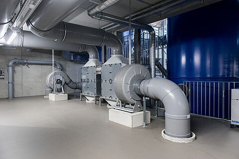 Ein technischer Raum mit großen Lüftungsrohren und mehreren Ventilatoren. Die Wände sind aus Beton, während eine Wand blau gestrichen ist. Auf dem Boden sind keine sichtbaren Objekte, und die Umgebung wirkt industriell.