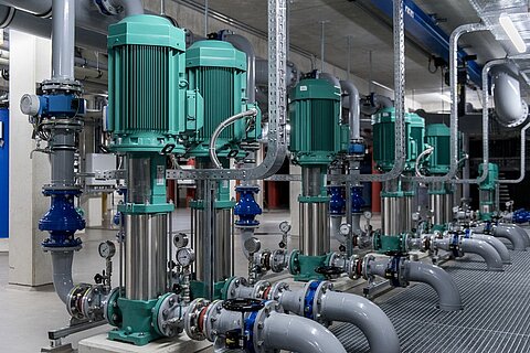 Eine Reihe von grünen Pumpen mit Edelstahlgehäusen ist in einem Industriegebäude angeordnet. Die Pumpen sind an Rohre und Armaturen angeschlossen, die aus grauem Material bestehen. Die Umgebung wirkt modern und technisch, mit sichtbar verlegten Rohrleitungen und Ventilen.