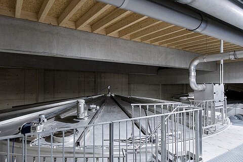 Eine industrielle Umgebung mit großen, runden Tanks und Metallrohren. Der Boden ist aus Beton, und die Decke ist aus Holz mit sichtbaren Balken. Die Tanks sind mit einem Geländer umgeben und scheinen Teil eines Produktions- oder Lagerprozesses zu sein.