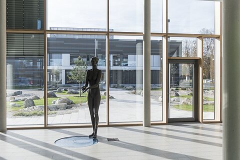 Im Innenraum eines modernen Gebäudes steht eine Skulptur in menschlicher Form vor großen Fenstern. Draußen ist ein gepflegter Garten mit Steinen und Bäumen sichtbar. Das Licht fällt sanft auf den Boden.