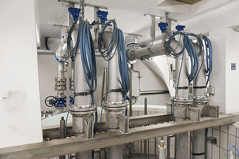 Das Bild zeigt mehrere große, intricate Rohrleitungen aus Metall, die durch einen Raum verlaufen. Einige Rohre sind mit blauen Schläuchen verbunden, und es sind verschiedene Ventile und Armaturen sichtbar. Der Raum hat weiße Wände und eine helle Decke.