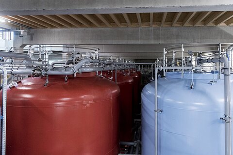 Mehrere große Lagertanks in rot und blau stehen in einem Industriegebäude. Rohrleitungssysteme sind sichtbar, die die Tanks miteinander verbinden. Das Licht fällt von oben auf die Tanks, die in einem modernen, strukturierten Raum angeordnet sind.