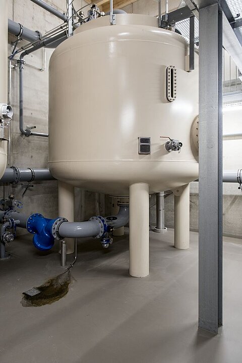 Großer, runder Tank in einer industriellen Umgebung. Der Tank ist hellbeige und steht auf vier Beinen. Rohrleitungen führen zu und von dem Tank, wobei ein blauer Armatur sichtbar ist. Der Boden ist grau und leicht nass.