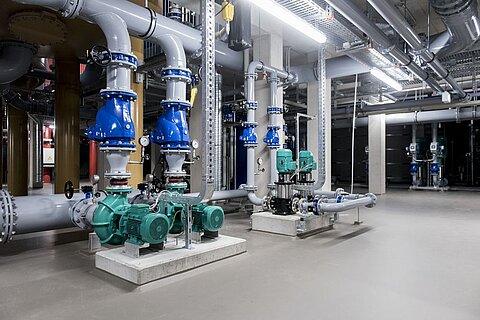 Ein industrieller Raum mit Rohrleitungen, Pumpen und Ventilen. Sichtbare grüne Elektromotoren und blaue Pumpen sind in einer sauberen Umgebung angeordnet, die für Versorgungssysteme genutzt wird. Die Beleuchtung ist hell und die Wände sind neutral gehalten.