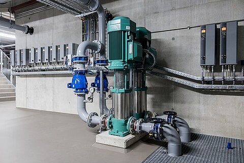 Das Bild zeigt eine industrielle Pumpanlage mit einer großen grünen Pumpe, die an Rohre und Ventile angeschlossen ist. Im Hintergrund befinden sich mehrere elektrische Steuergeräte an einer Betonwand. Der Boden ist aus grauem Material und die Umgebung wirkt funktional und technisch.