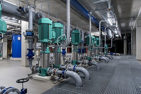 Eine Reihe von grünen Pumpen und Rohrleitungen in einem modernen, industriellen Raum. Der Boden ist mit Gitterfliesen ausgestattet, und es sind verschiedene Ventile und Armaturen sichtbar. Die Umgebung wirkt sauber und gut organisiert, mit sichtbaren Leitungen an der Decke.