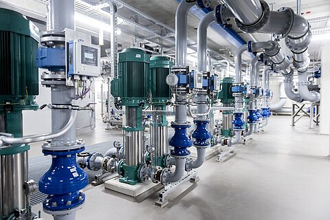 Eine Reihe von industriellen Pumpen und Rohren in einem modernen technischen Raum. Die grünen Pumpen stehen an einer Wand, verbunden mit blauen Ventilen und verschiedenen Messgeräten. Der Boden ist grau und die Beleuchtung ist hell.