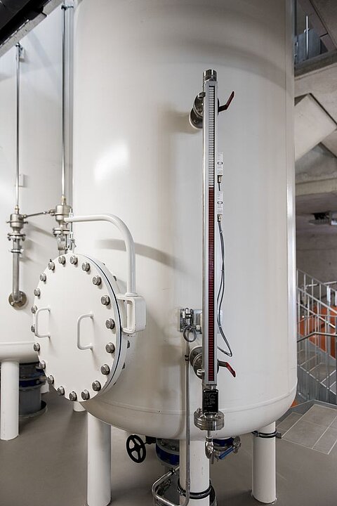 Ein großer, weißer Edelstahlbehälter steht in einem industriellen Raum. An der Seite sind Rohre und ein Messinstrument angebracht. Der Behälter hat eine ausgeprägte runde Tür mit Schrauben und befindet sich auf einem grauen Fliesenboden in der Nähe einer Treppe.
